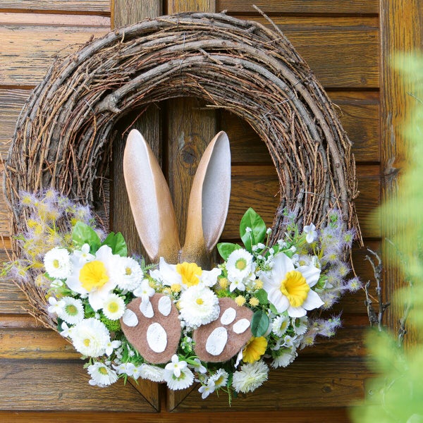 Osterkranz aus Weidenzweigen geschmückt mit Osterhasenohren, Pfoten und Frühlingsblumen wie Narzissen, hängend an einer Holztür.