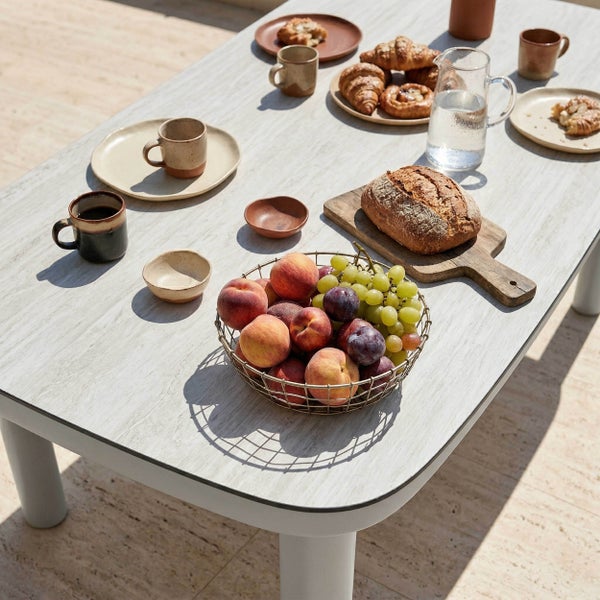 Moderner Gartentisch mit Keramikplatte, gedeckt mit Obstschale, Brot auf Holzbrett und Keramikgeschirr im hellen Außenbereich.