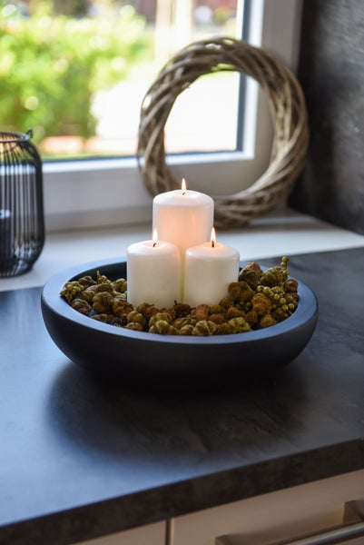 Drei brennende Stumpenkerzen in einer flachen Schale mit dekorativen Naturmaterialien auf einer dunklen Arbeitsplatte vor einem hellen Fenster.
