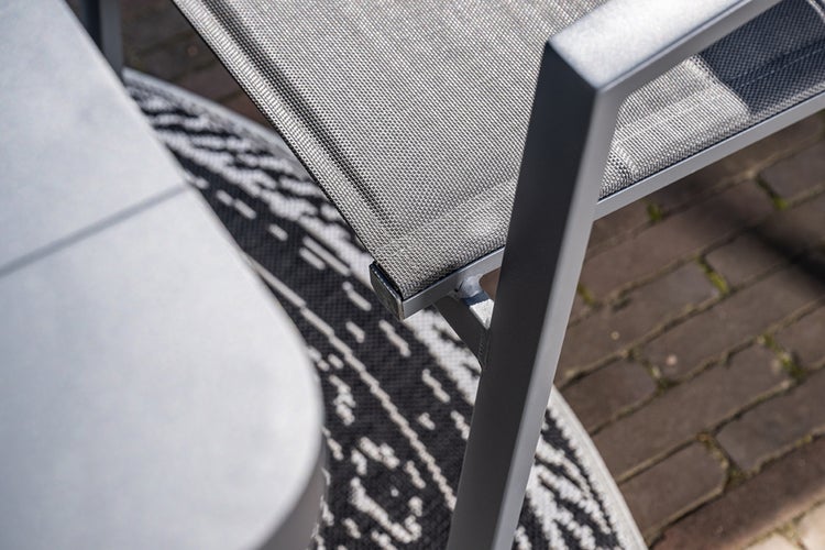 Detailansicht eines Gartenstuhls mit Aluminiumrahmen und Netzgewebe neben einem Gartentisch auf einem Außenteppich.