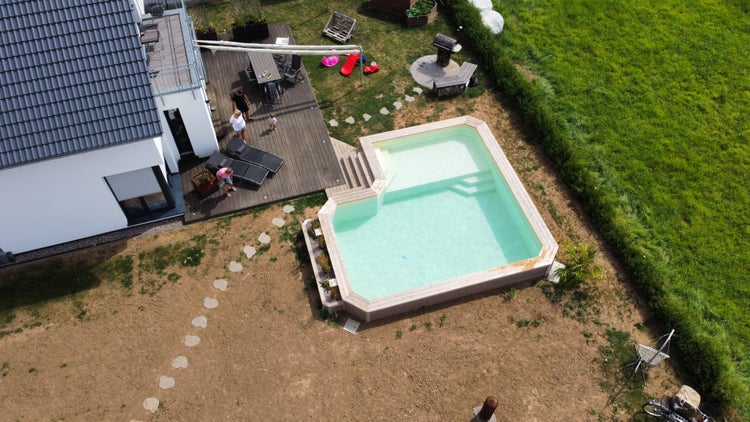 Luftaufnahme eines L-förmigen Swimmingpools mit Holzumrandung in einem Garten neben einem Wohnhaus mit Holzterrasse.
