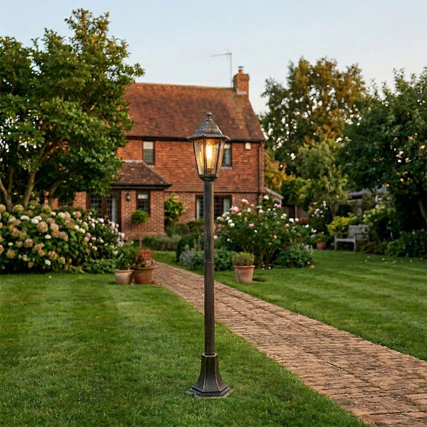 Klassische Gartenlaterne auf grünem Rasen neben einem Backsteinweg, im Hintergrund ein Garten mit Blumen und einem Backsteinhaus.