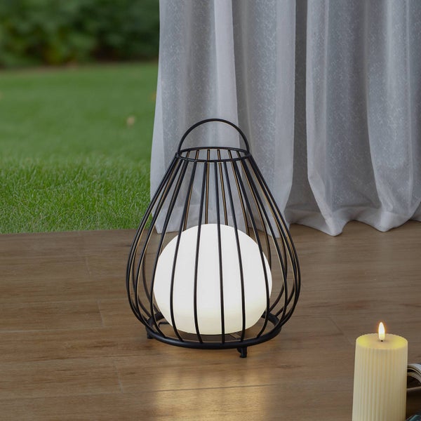 Dekorative schwarze Käfig-Laterne mit leuchtender Kugel auf einer Holzterrasse vor einem weißen Vorhang und Rasen.