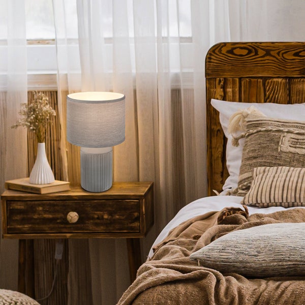 Tischleuchte mit grauem Stoffschirm und geriffeltem Keramikfuß auf einem hölzernen Nachttisch in einem hellen, rustikalen Schlafzimmer.