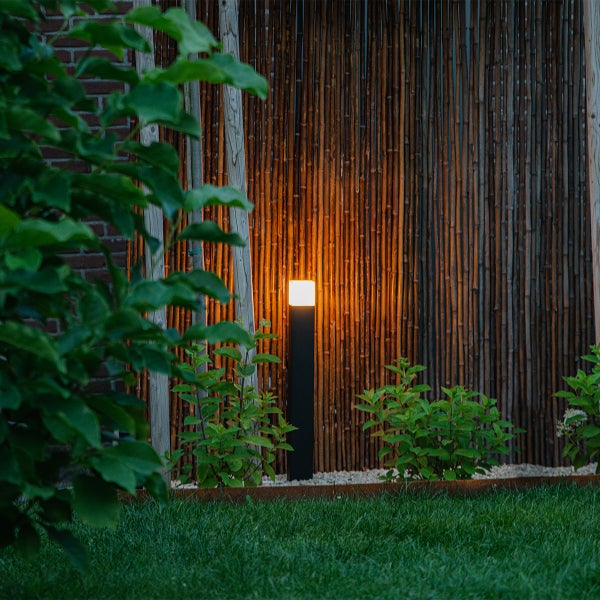 Moderne schwarze Wegeleuchte mit warmem Licht vor einer Bambuswand in einem Garten mit Rasen und Pflanzen.