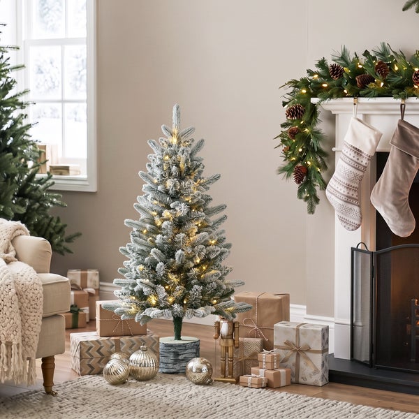 Weihnachtliche Szene mit beleuchtetem, künstlichem Weihnachtsbaum, der mit Schnee bedeckt ist, vor einem Kamin mit Girlande und Weihnachtsgeschenken.