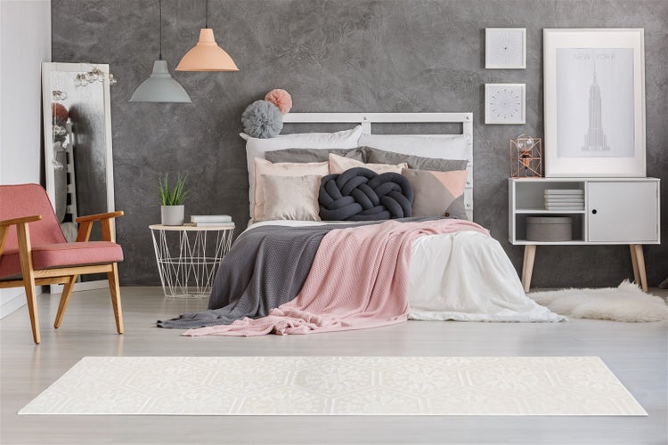 Ein moderner heller Teppich mit hexagonalem Muster liegt vor einem stilvoll eingerichteten Schlafzimmer mit grauer Wand und weichen Textilien.