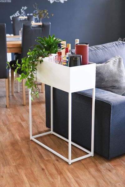 Weißer Pflanzkasten aus Metall auf hohem Gestell, genutzt als Beistelltisch für Pflanzen und Bar-Zubehör neben einem Sofa.