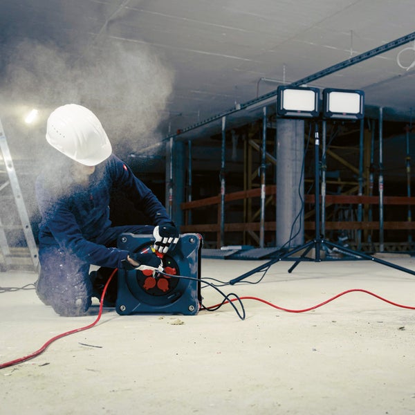 Ein Bauarbeiter mit Helm arbeitet mit einer Kabeltrommel auf einer Baustelle, im Hintergrund Strahler.