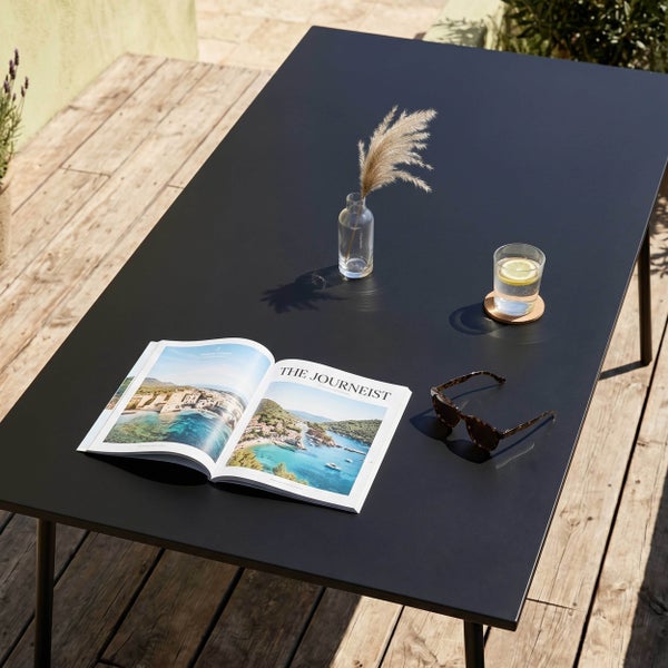 Rechteckiger Gartentisch aus Metall auf einer Holzterrasse mit einer Zeitschrift, Sonnenbrille und einem Glas Wasser.