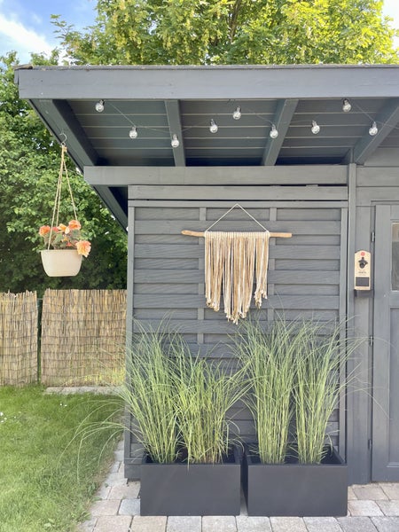 Graues Gartenhaus mit Lichterkette, Makramee Wandbehang, Blumenampel und schwarzen Pflanzkübeln mit Ziergras vor einem Bambus Sichtschutz im Garten.