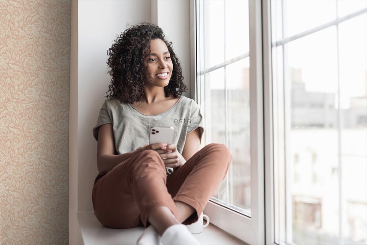 Eine Frau sitzt am Fenster und blickt nach draußen, während sie ein Smartphone in der Hand hält. Die Wand ist mit einer floralen Tapete verziert.