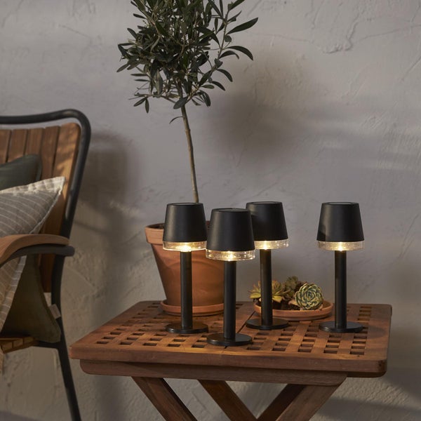 Vier kleine, schwarze Tischleuchten auf einem Holztisch mit Pflanze