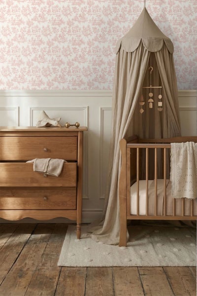 Klassisch eingerichtetes Kinderzimmer mit Mustertapete, Wandtäfelung, einer Holzkommode und einem Babybett mit Stoffbaldachin.