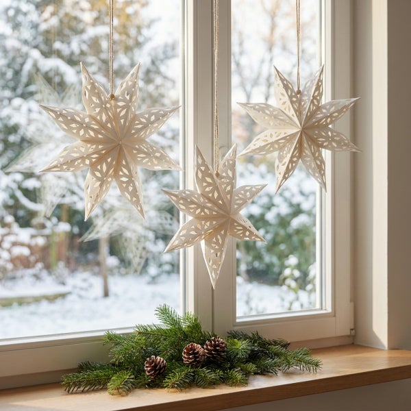 Drei hängende Papiersterne zur Fensterdekoration vor einem verschneiten Garten mit Tannenzweigen und Zapfen auf der Fensterbank.