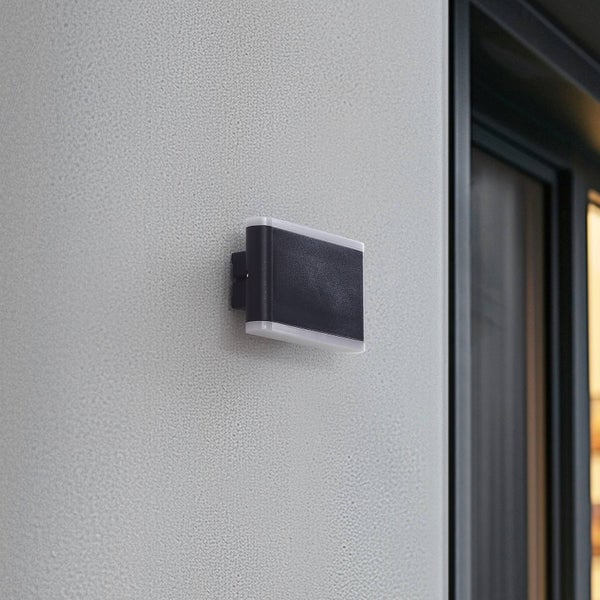 Moderne rechteckige Außenwandleuchte an einer hellen Putzfassade neben einem dunklen Türrahmen.