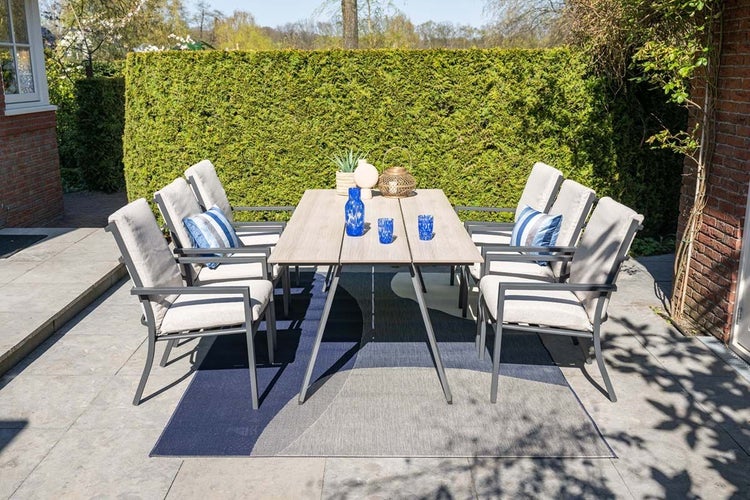 Moderne Gartengarnitur mit einem rechteckigen Tisch in Holzoptik und sechs gepolsterten Sesseln auf einer hellen Terrasse vor einer grünen Hecke.