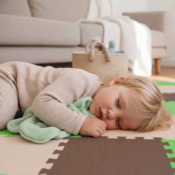 Ein Kleinkind schläft auf einer Puzzlematte aus Schaumstoff mit einem Kuscheltuch, im Hintergrund steht ein Korb im Wohnzimmer.