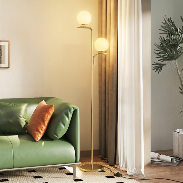 Moderne Stehleuchte mit goldfarbenem Metallgestell und zwei weißen Glaskugeln in einem hellen Wohnzimmer mit grünem Sofa, Vorhängen und einer Pflanze.