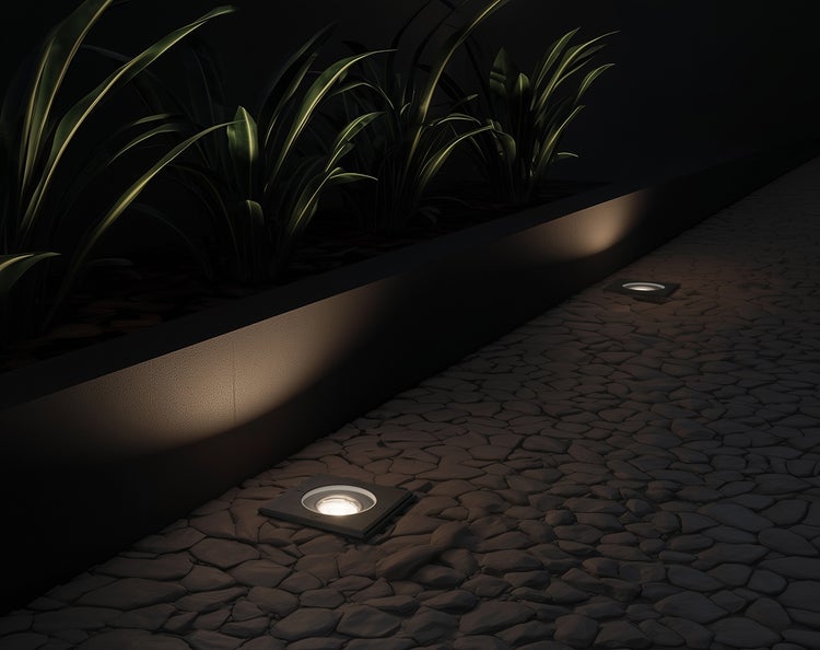 Einbaustrahler im Außenbereich beleuchten einen Gartenweg aus dunklen Pflastersteinen neben einem Pflanzenbeet bei Nacht.