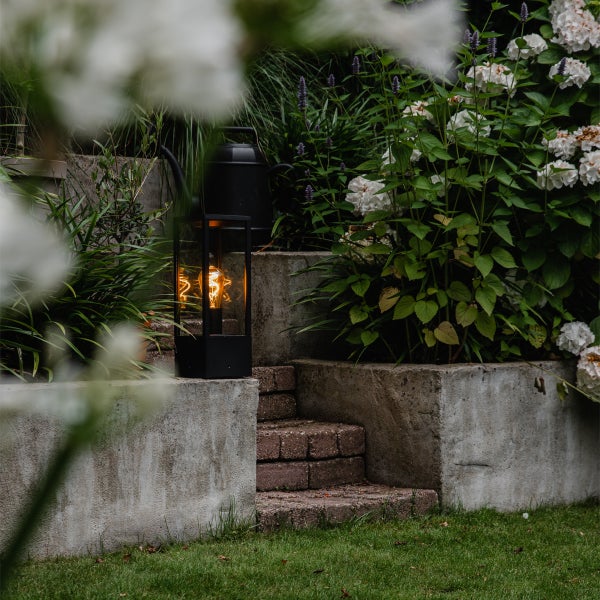 Eine schwarze Außenlaterne mit dekorativer Leuchtwendel steht auf einer Gartenmauer aus Stein inmitten von Hortensien und Grünpflanzen.