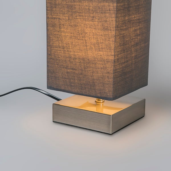 Eckige Tischleuchte mit Metallfuß und dunklem Stoffschirm, eingeschaltet mit Netzkabel.