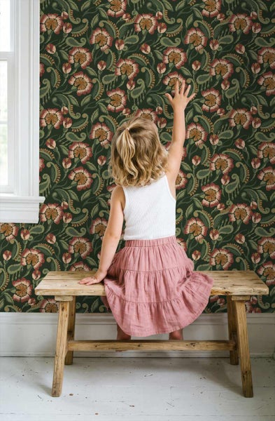 Vliestapete mit floralem Paisleymuster in Dunkelgrün, Rot und Beige. Ein Kind sitzt auf einer rustikalen Holzbank vor der tapezierten Wand.