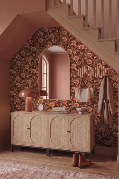 Organisierter Eingangsbereich unter einer Holztreppe mit Blumentapete, Sideboard mit Geflecht-Türen, Rundbogenspiegel und Garderobe.