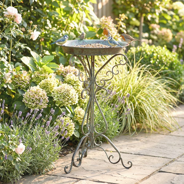 Dekorative Vogeltränke aus Metall auf einem verzierten Standfuß in einem hellen Garten mit Blumen.