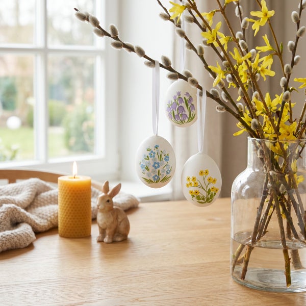 Osterdekoration mit Weidenkätzchenzweigen, hängenden Ostereiern mit Blumenmotiven, einer Kerze und einer Hasenfigur auf einem Holztisch.