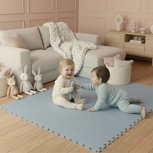 Zwei Babys spielen auf einer hellblauen Puzzle-Spielmatte im hellen Wohnzimmer mit Sofa, Holzboden und Wandpaneelen.
