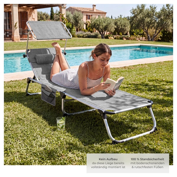 Eine Frau liegt auf einer Gartenliege mit Sonnendach und Kopfkissen auf einer Wiese neben einem Pool und liest ein Buch.