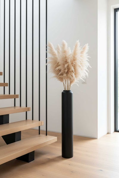 Hohe, schwarze Bodenvase mit Pampasgras neben einer Holztreppe mit schwarzen Metallstäben in einem hellen Raum.