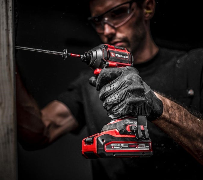 Person mit Schutzbrille und Handschuhen nutzt einen Einhell Akku-Schlagschrauber Power X-Change, um eine Schraube in Holz zu drehen.