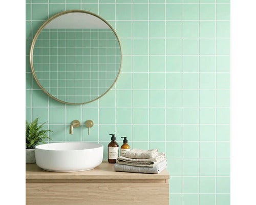 Helles Badezimmer mit mintgrünen Wandfliesen, rundem Spiegel mit Goldrahmen, weißem Aufsatzwaschbecken und goldener Armatur auf Holzunterschrank.