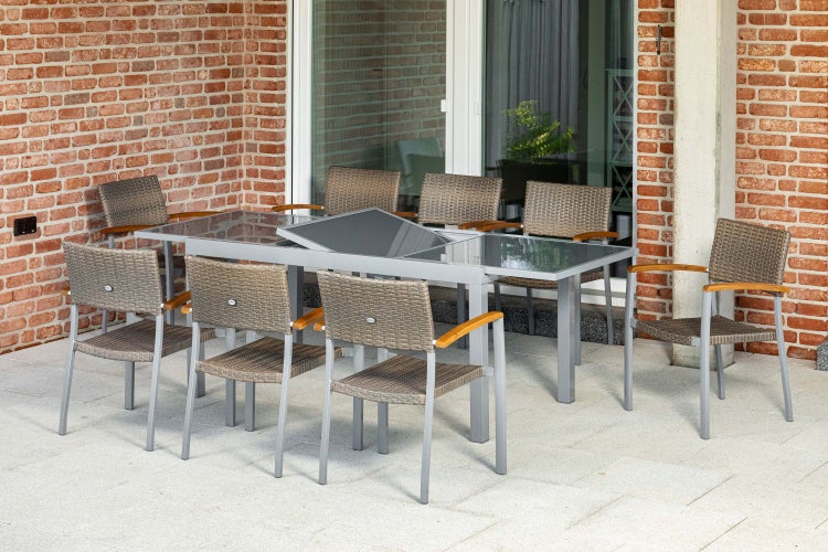Gartengarnitur mit ausziehbarem Tisch mit Glasplatte und acht Stühlen mit Geflecht und Holzarmlehnen auf einer Terrasse vor einer Backsteinwand.