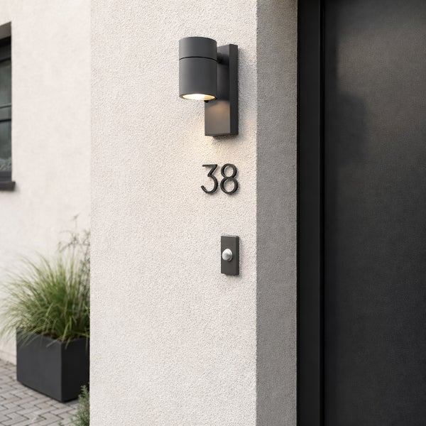 Moderne zylindrische Außenwandleuchte mit Hausnummer 38 und Klingeltaster an einer hellen Fassade neben einer dunklen Haustür.