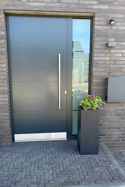 Moderne Eingangssituation mit anthrazitfarbener Haustür, langem Edelstahlgriff, Glasseitenteil und hohem Pflanzgefäß vor einer Ziegelwand.
