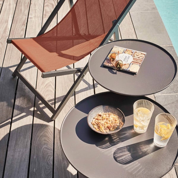 Moderne Terrassenszene mit rostfarbenem Liegestuhl auf Holzdeck neben Pool und zwei schwarzen runden Beistelltischen mit Erfrischungen.