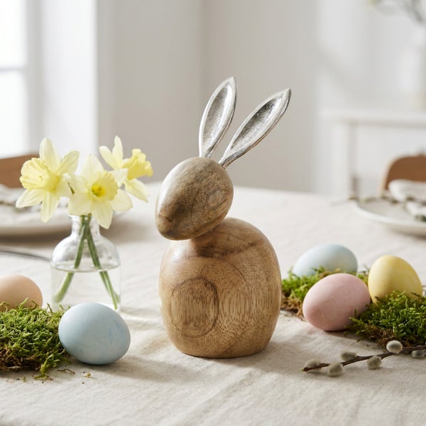 Osterdekoration mit einem Osterhasen aus Holz mit Metallohren, umgeben von Ostereiern, Frühlingsblumen und Weidenkätzchen auf einem Tisch.