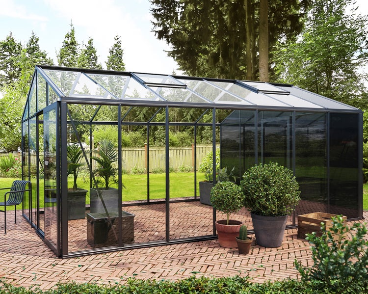 Modernes Gewächshaus aus Glas mit anthrazitfarbenem Aluminiumrahmen auf einer gepflasterten Terrasse im Garten mit verschiedenen Pflanzen.