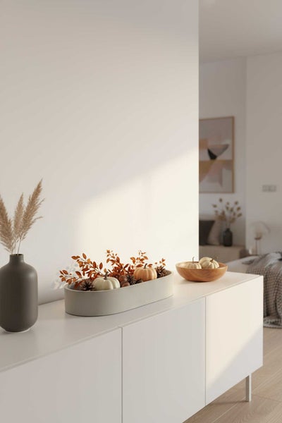 Moderne Herbstdekoration auf einem weißen Sideboard mit Vase, dekorativer Schale mit Kürbissen und Zapfen in einem hellen Raum.