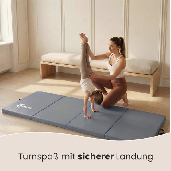 Eine Frau hilft einem Kind beim Handstand auf einer grauen Turnmatte.