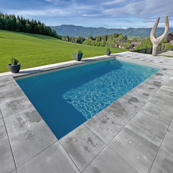 Rechteckiger Außenpool mit blauem Wasser, grauen Steinterrassenplatten und Pflanzkübeln in einer weitläufigen Hügel- und Gebirgslandschaft.