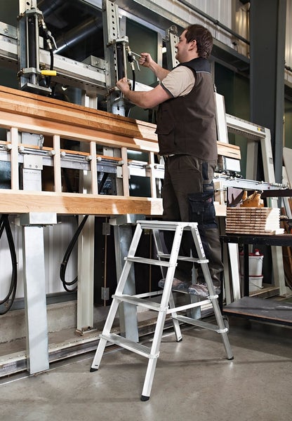 Handwerker steht auf einer Aluminium Trittleiter und bedient eine Holzpresse in einer Werkstatt.