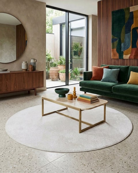 Modern eingerichtetes Wohnzimmer mit hellem runden Teppich, grünem Sofa, Couchtisch aus Stein und Holz-Wandpaneelen vor Fensterfront.