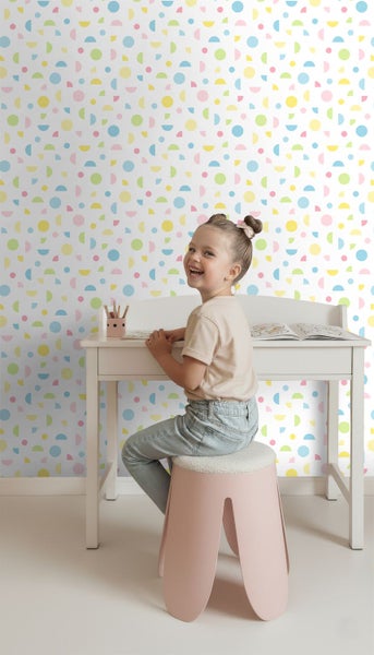 Helles Kinderzimmer mit gemusterter Tapete mit bunten geometrischen Kreisen und Halbkreisen, ein Mädchen sitzt an einem weißen Schreibtisch.