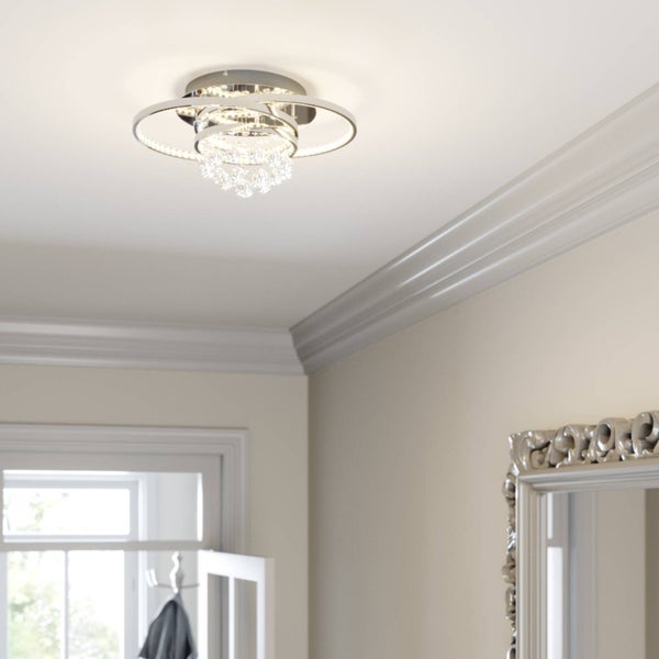 Moderne LED-Deckenleuchte mit ineinander verschlungenen Chromringen und Kristallanhängern in einem hellen Raum mit Stuckleisten.