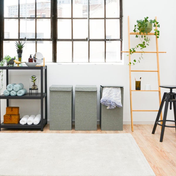 Heller, organisierter Raum mit drei grauen Wäschekörben aus Stoff, einem schwarzen Metallregal, einer Holzleiter, einem Hocker und großem Fenster.