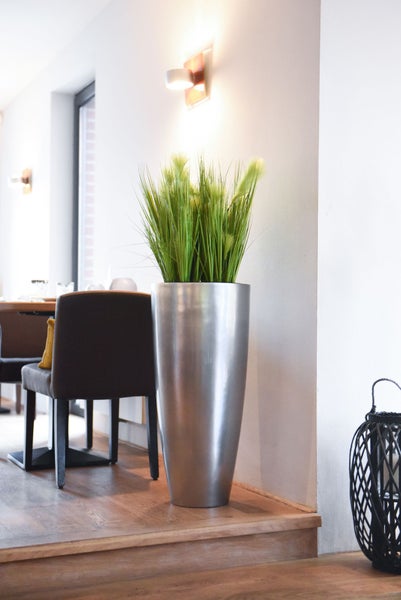 Hohes silbernes Pflanzgefäß aus Metall mit künstlichem Gras in einem modernen, hellen Raum mit Holzboden und dunklem Stuhl.